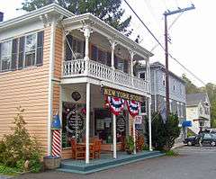An orange building with a white porch and a storefront at street level with red, white and blue bunting and a sign saying "New York Store". Farther down the side of the street are a gray building and a yellow one.