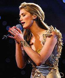 A female with light brown hair singing into a microphone, while holding the microphone with her two hands. The female is wearing a bracelet on her left arm and red nail polish on both her hands finger nails.