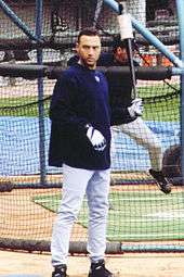 Derek Jeter stands in front of a batting cage wearing a navy long-sleeve shirt over a baseball uniform while holding a weighted bat in his left hand.