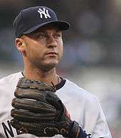 Derek Jeter wearing a navy hat and grey baseball uniform with a black glove stares into the distance.