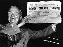 Man in gray suit and wire glasses holding newspaper that says "Dewey Defeats Truman"