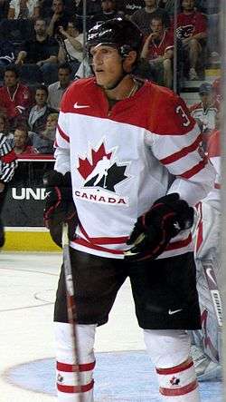 Hockey player in white Canada uniform. His hands are by his side, one holding his stick, and he stands near the sideline of the rink.