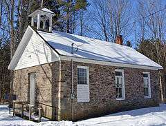 A small stone building viewed from the side in winter. Snow covers the ground and its black roof, which has a small empty steeple and a brick chimney