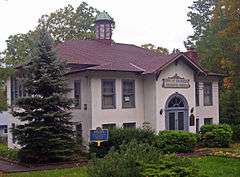 A tan building with brown roof and a cupola on its peaked roof and pale blue window and door trim. Shrubbery is in front with taller trees in back, and there is a blue and gold historical marker in front saying "1925: School House District 14 Has Been placed on the National Register of Historic Places 1997"