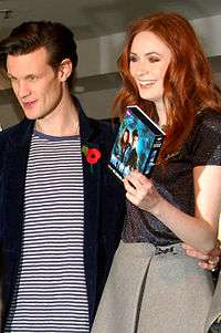 Matt Smith and Karen Gillan are turned to the side and smiling as if taking a picture. Gillan is holding up a DVD boxset of the series.