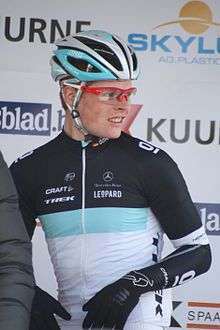 A man in his mid-twenties wearing a cycling jersey with black sleeves, a white torso, and a blue stripe separating them, along with red-framed glasses. He is looking to his left off-frame.