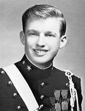 A black-and-white photograph of Donald Trump as a teenager, smiling and wearing a dark uniform with various badges and a light-colored stripe crossing his right shoulder. This image was taken while Trump was in the New York Military Academy in 1964.