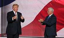 Donald Trump and his running mate for vice president, Mike Pence, at the Republican National Convention in July 2016. They appear to be standing in front of a huge screen with the colors of the American flag displayed on it. Trump is at left, facing toward the viewer and making "thumbs-up" gestures with both hands. Pence is at right, facing toward Trump and clapping.