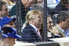 Trump at a baseball game in 2009. He is wearing a baseball cap and sitting amid a large crowd, behind a protective net.