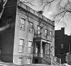 A black and white photograph showing a brick house from its front right. There is some remaining snow on the ground, and the top is partially obstructed by bare tree branches. The front of the house has an ornate portico with columns and a flat roof.