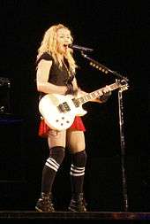 Madonna in red shorts and black T-shirt playing a white electric guitar.