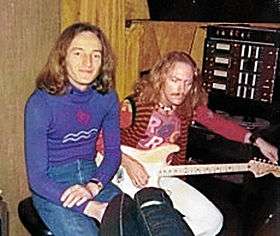 Two musicians sit side by side in a recording studio. Both men have long red hair. The man on the left wears a blue jumper and sits in a relaxed manner. The one on the right is tuning his guitar.