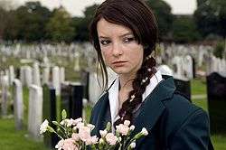 A teenage girl wearing a school uniform stands in a graveyard, holding flowers