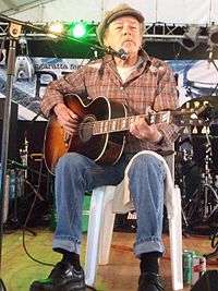 A sixty-nine-year old man is playing a guitar. He is sitting on a white plastic chair, strumming with his right hand while staring to his left. A microphone with its stand is near his chin. He wears a cap, check shirt, blue jeans and black shoes. Behind him is stage equipment, at his feet to the left of his chair are three cans of drink.