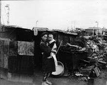 Black and white photograph of a woman wearing traditional Japanese clothing holding a small child while standing in front of a crudely built shack. Rubble and undamaged houses are visible in the background.