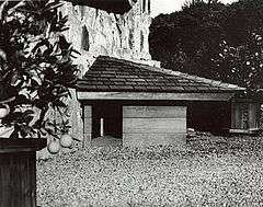 A black and white photo of a small triangular doghouse with a door on its left