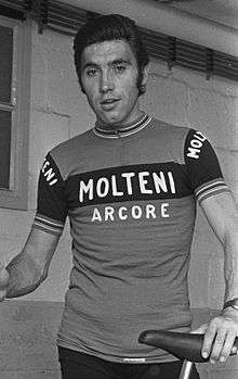 Merckx holding a bicycle. His shirt says "Molteni Arcore", and his hair is slicked back.