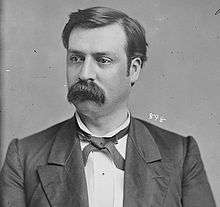 A man with dark hair and a mustache wearing a dark jacket and tie and white shirt