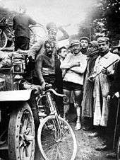On the left a classic automobile, and a group of men standing, one of them holding a bicycle.