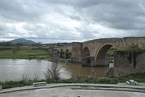 Photo of an old arched stone bridge over a river. There is a mountain in the distance