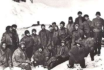  A group of men sitting closely packed together, in heavy winter clothes and wearing hats. Snow and ice on the ground and in the background