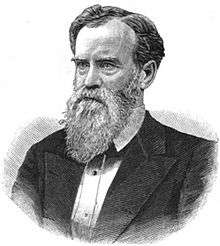 A man with black hair and a graying beard wearing a black jacket and white shirt