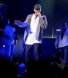 A man on a stage wearing blue jeans, a white shirt, a grey jacket and a cap; two men can be seen in the background