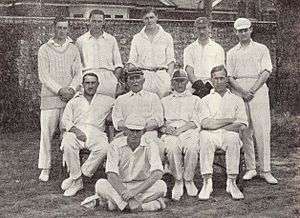 A cricket team arranged in two rows. Most are wearing cricket whites and caps.