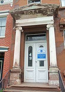 A yellowed, damaged wooden pediment supported by fluted columns with Ionic capitals sheltering an entrance with a large "49" on one white door and a sign listing a phone number on another