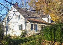 A stone house painted white seen from the left side with the front visible. There are shrubs in front and some trees with yellow leaves in back.