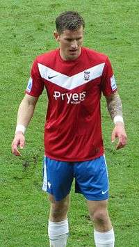 A man with brown bhair who is wearing a red top, blue shorts and white socks. He is standing on a grass field.