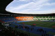 An oval shaped stadium with a blue athletics track and stands with red, blue, orange and green seating sections. One of the lateral stands is filled and more people are in the track. The sky is blue with clouds.