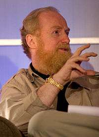 Action portrait of a man in his fifties with a bushy, strawberry-blond beard seated while speaking wearing a safari jacket and gold watch