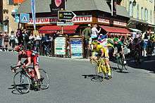 A line of cyclists, led by one in a black and red jersey with white trim who is followed by Voeckler in his all-yellow jersey. More cyclists follow, and spectators look on from the roadside.