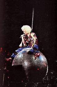 Image shows female performer sitting on a disco ball, wearing a big curly blonde wig, a maroon blouse and blue pants with red heels and is holding a microphone to her mouth.