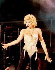The image of a young blond woman. She is wearing a beige corset and black pants. She has blonde curly hair and has a headset microphone to her mouth.