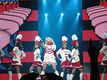 Picture of several women wearing white and red majorette uniforms. The one in the middle is blonde and is holding a baton in front of her. The backdrops show a red and white animation