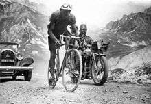 A cyclist climbing a mountain, followed by a motorcycle and a car.