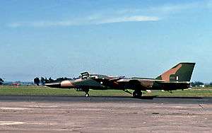 Jet aircraft parked on ramp perpendicular to camera facing left. It wears a green and brown camouflage scheme.
