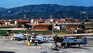Colour photograph of an airfield. Three single-seat piston-engined propeller aircraft sit in the foreground, as a number of personnel work on the machines which are finished in natural metal with blue, white and red roundels. In the middle distance are a number of makeshift corrogated-iron buildings, while to their rear are several others of more solid construction. In the background rise a number of tree covered mountains.