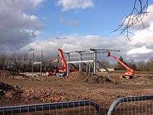 A muddy construction site with two vehicles working to build the stadium during the day. A small steel structure has been created which is beginning to resemble a football stand. The sky is lightly cloudy and in the background there are trees.