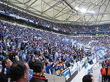 A pitch level perspective of the corner section of a stadium filled with people holding blue scarfs and waving blue-and-white flags.