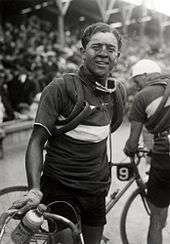 A man with a dirty face holding his bicycle, spare tires wrapped around his shoulders. In the background a large crowd.