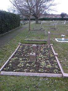 A flat grave surrounded by a stone border with small plants in tended bare earth. In the centre are two stone blocks. In the background are similar grass-covered graves.