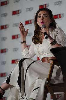Photograph of a brunette white woman sitting on a chair and holding a microphone.