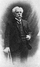 Portrait photograph of a standing man with white hair and large white moustache, wearing a dark three-piece suit