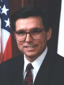 Hispanic man with large glasses and black hair with the US flag behind him