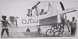 Two men seated in a World War I-era biplane, surrounded by technical personnel