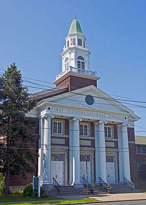 A two-story brick building with a white colonnade on front, topped by a green rounded cupola, in the American Colonial Revival style. "First Congregational Church" is engraved across the front above the columns. It is seen from across the street, with wires in front of it.