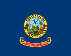 A blue flag with a circular seal in the center. The words "State of Idaho" appear in gold letters on a red and gold band below the seal.